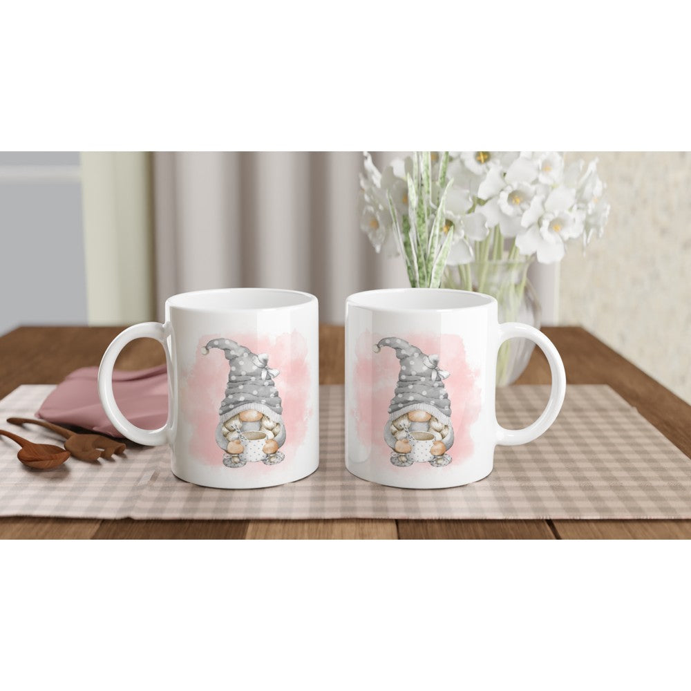 Two Pink and Grey Gonk Mugs sit on a light-colored checkered placemat on a wooden table. Behind the mugs, there's a white vase with white flowers and a pink envelope beside wooden utensils. The bright indoor setting perfectly complements this charming home decor.
