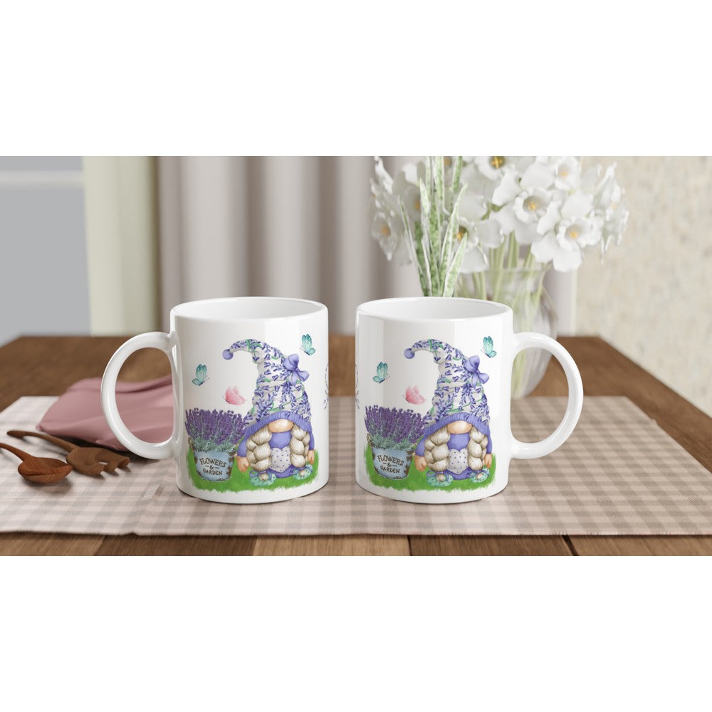 Two Gonk Lavender & Butterfly Mugs with matching whimsical gnome illustrations, butterflies, and lavender plants are placed side by side on a checkered tablecloth. White flowers in the background and wooden kitchen utensils on the left complete the cozy home decor setting.